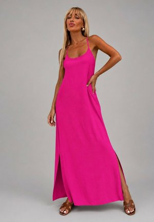 Femme aux longs cheveux blonds portant une robe maxi rose vif sans manches avec des fentes latérales et des sandales marron, debout devant un fond gris.