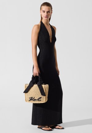 Kobieta w czarnej, bezrękawnikowej, długiej sukni z wiązaniem na szyi, trzymająca plecioną beżową torebkę z czarnymi wstążkowymi uchwytami, stojąca na szarym tle.