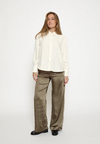 Kreemikaalusega pikkade varrukatega nööbitav särk koos laia säärega läikivate oliivroheline pükstega. Mustad kingad täiustavad komplekti.