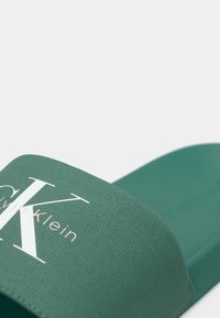 Infradito verde con cinturino in tessuto testurizzato e plantare liscio. Presenta un logo bianco prominente e un design minimalista.