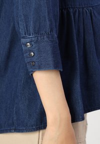 Dark blue denim shirt with a gathered waist, buttoned cuffs, and a smooth texture. Features four dark buttons on the sleeve for closure.