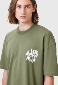 Olchowa zielona, oversize'owa koszulka bawełniana z białym graficznym logo na lewej klatce piersiowej. Krótkie rękawy i dekolt okrągły.
