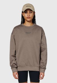 Brun sweatshirt med ribbade manschetter och kant, med "HALO"-logotyp i grönt. Ihop med en beige baseballkeps och svarta shorts.