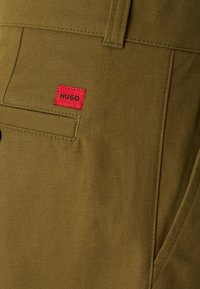 Detailný záber na vrecko olivovo zelených nohavíc s červeným obdĺžnikovým štítkom s nápisom "HUGO" v čiernom texte nad otvorom vrecka.
