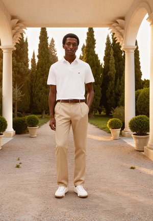 Un homme se tient sur un chemin de gravier sous une arche, portant un polo blanc, un pantalon beige, des baskets blanches, entouré de plantes en pot et de grands arbres verts.
