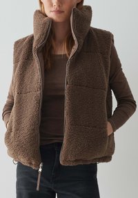 Brauner Fleece-Weste mit hohem Kragen, Reißverschluss und seitlichen Taschen. Hat eine strukturierte Oberfläche und ein glattes Innenfutter.