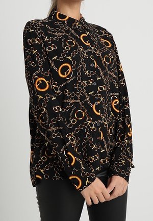 Kvinde iført en sort skjorte med knapper med et mønster af orange og guld kæder og bælte, kombineret med sorte bukser, står med hænderne foldet.