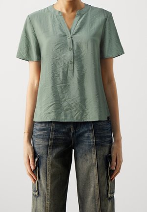 Woman wearing a wrinkled sage green short-sleeve V-neck blouse and faded blue denim cargo pants, standing against a plain background.