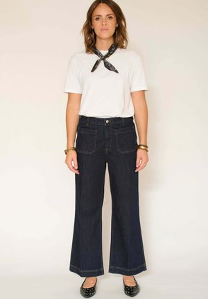 Jeans en denim foncé à jambes larges avec poches avant et arrière, associés à un t-shirt blanc à col rond et un foulard noir à motifs autour du cou. Des ballerines noires sont portées.