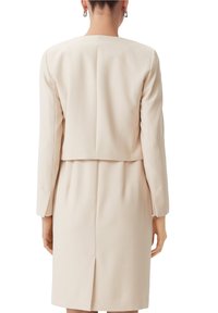 Beige klänning med struktur, långärmad och med en kort ryggdesign. Har en slät textur och en central söm baktill.