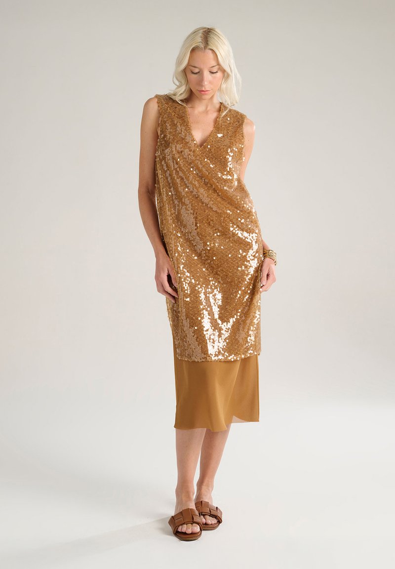 Abito dorato con paillettes, scollatura a V e design drappeggiato, sovrapposto a un abito sottile di raso marrone. Modella che indossa sandali scivolosi marroni.
