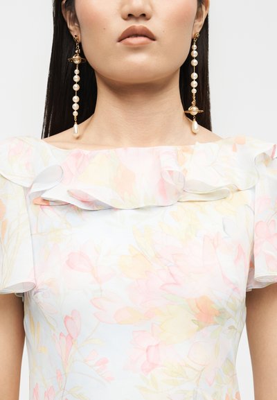 Γυναίκα που φορά μακριά χρυσά και μαργαριταρένια κρεμαστά σκουλαρίκια και παστέλ φορέματα με φλοράλ σχέδιο και γιακά με βολάν.