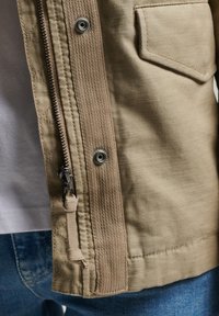 Khaki jakke med lynlås i metal, trykknap og en lommepatch. Stoffet er tekstureret med en vævet detalje langs lynlåsen.