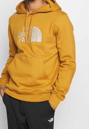 Person iført en karrygul hættetrøje med "The North Face"-logo og sorte bukser med logo på venstre lår, hænderne i lommerne.