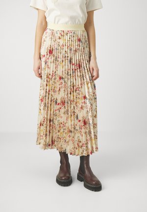 Kvinde iført en plisseret beige floral midi-nederdel kombineret med mørkebrune chunky Chelsea-støvler, stående op ad en ensfarvet baggrund.
