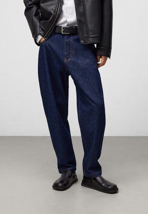 Jeans in denim blu scuro con una vestibilità ampia, accentuati da cuciture arancioni. Abbinati a scarpe di pelle nera e una giacca di pelle nera.