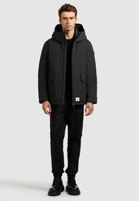 Schwarze gepolsterte Jacke mit Kapuze, Reißverschluss vorne und zwei Vordertaschen. Wird über einem schwarzen Hemd getragen und kombiniert mit schwarzen Cargo-Hosen und Stiefeln.