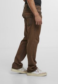 Pantalones de mezclilla marrón con costuras en contraste, ajuste relajado y diseño de cinco bolsillos. Presenta una etiqueta de cuero en la parte posterior y un desgaste mínimo.