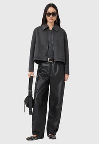 Veste grise coupe courte avec deux poches avant, associée à un pantalon en cuir noir ample et des ballerines décorées de clous. Modèle tenant un sac.