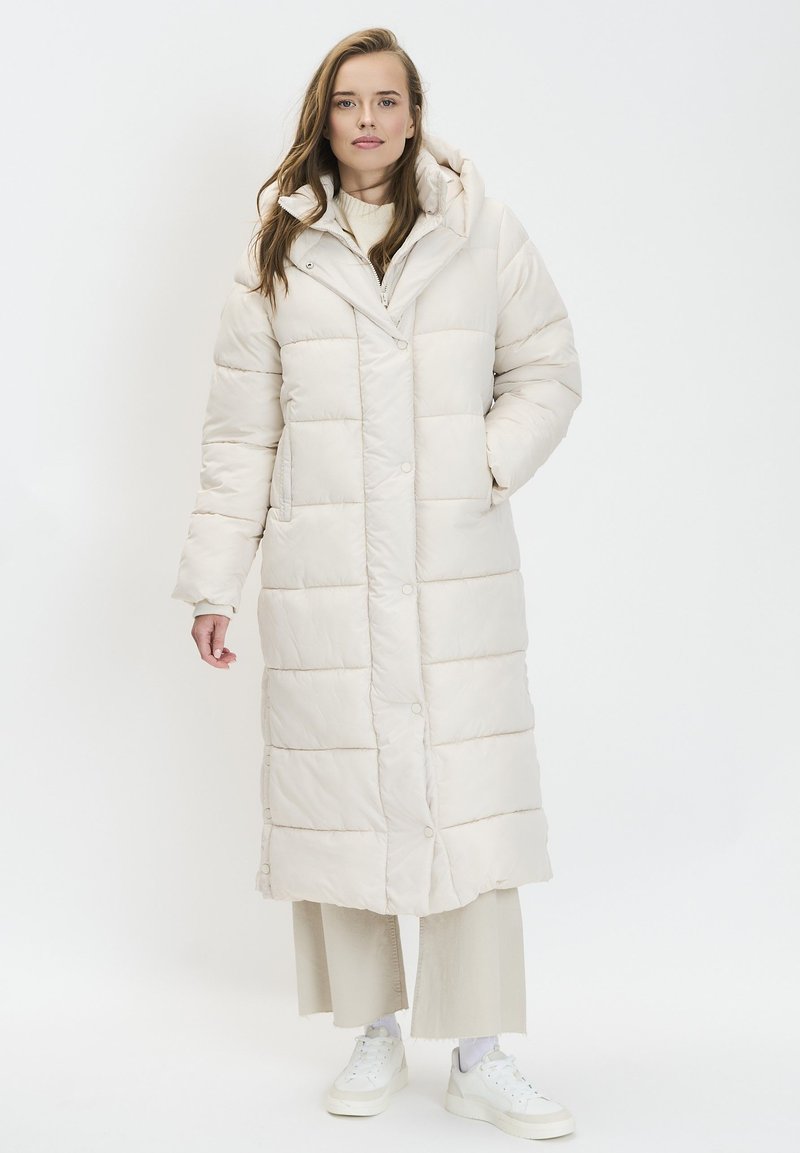 Manteau long matelassé beige clair avec capuche, doté d'une fermeture à boutons et de poches latérales. Texture lisse avec un design matelassé.