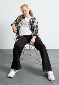 Pantalones de pierna ancha negros, camiseta gráfica blanca y camisa de franela a cuadros. Modelo sentado en una silla de alambre, usando zapatillas blancas.