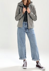 Veste grise avec capuche, dotée d'une fermeture éclair et de poches. Associée à un jean ample bleu clair et des baskets bordeaux foncé. Chemise rayée en dessous.