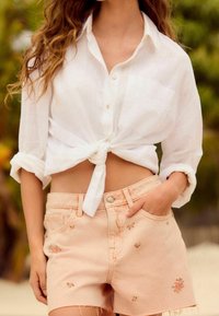 Camisa blanca de botones atada en la cintura; pantalones cortos de mezclilla color durazno con bordados florales y bordes deshilachados. Textura suave y ajuste casual.