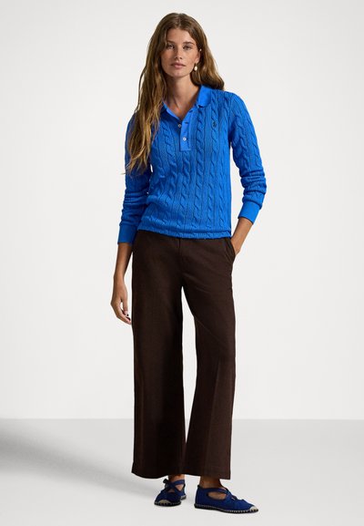 Moteris su ilgais plaukais, vilkinti ryškiai mėlyną megztinį polo stiliaus su virvutės raštu, tamsiai rudus plačiomis kojų kelnėmis ir derančius minkštus mėlynus batus, stovinti atsipalaidavusi.