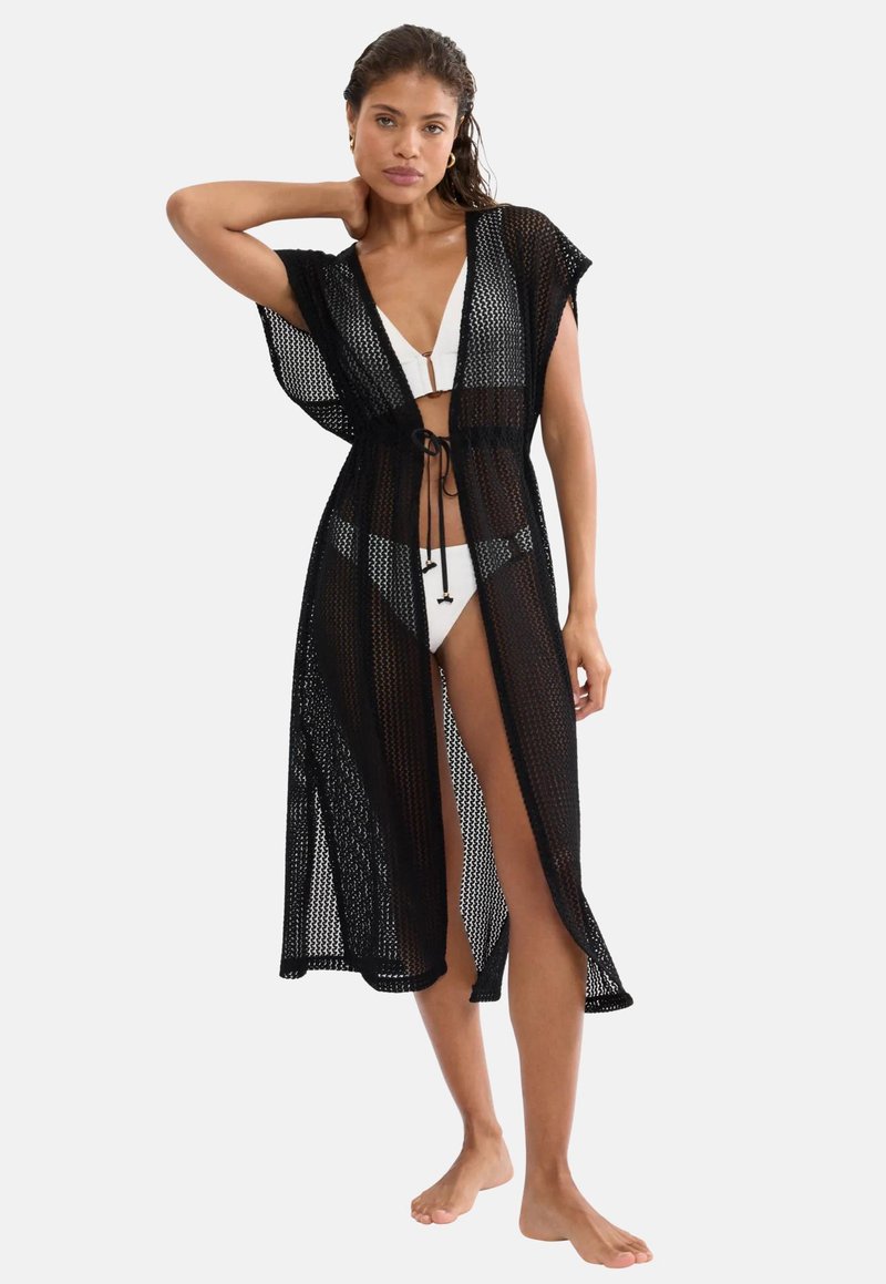 Γυναίκα που φοράει λευκό μπικίνι και μαύρο διαμπερή, μεσαίου μήκους κορδόνι με σούρα στη μέση, όρθια, ξυπόλυτη, μπροστά σε απλό φόντο.