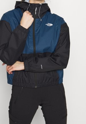 Person wearing a blue and black The North Face hooded jacket with a front zipper and black pants, posing with arms crossed.