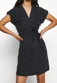 Vestito nero a portafoglio realizzato in tessuto leggero, con maniche corte, due tasche sul petto e un dettaglio in vita con fiocco.