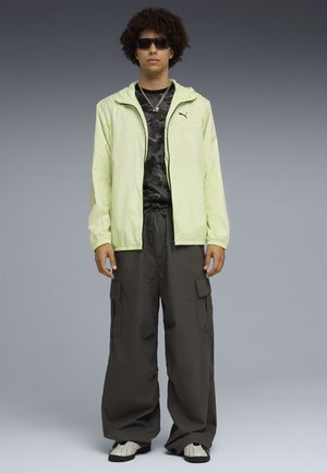 Jonge man met zwarte zonnebril, neon gele jas met rits, zwart camouflage shirt, losse donkere cargobroek, witte sneakers en zilveren kettingen, rechtopstaand.