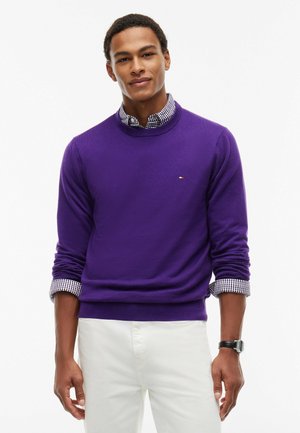 Giovane uomo che indossa un maglione viola sopra una camicia a quadri con le maniche rimboccate e pantaloni bianchi su uno sfondo semplice.