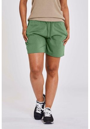 Person iført grønne shorts med snøre, beige top og sorte sneakers med hvide snørebånd, står med krydsede ben.