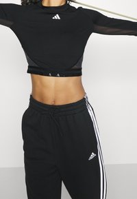 Žena v černém cropped topu Adidas s síťovými rukávy a černých teplácích Adidas s bílými pruhy, s vystrčenými pažemi.