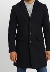 Cappotto in lana di un blu navy scuro con un design su misura, tre bottoni, rever a taglio e tasche laterali, indossato sopra un maglione grigio a trecce.