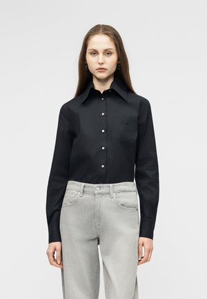 Νεαρή γυναίκα με μακριά ίσια μαλλιά, ντυμένη με μαύρο πουκάμισο με κουμπιά και ανοιχτό γκρι τζιν, στέκεται μπροστά σε απλό λευκό φόντο.