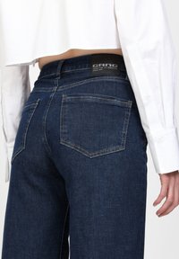 Dunkelblaue Jeans mit geradem Schnitt, mit einem schwarzen Etikett auf Höhe der Taille. Die Gesäßtaschen haben ein einfaches rechteckiges Design.
