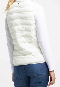 Vit, vadderad väst med horisontell söm, hög krage och en liten logga bak. Bärs över en långärmad topp och kombineras med jeans.