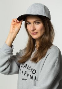Grå keps med en böjd skärm och textilstruktur, bärs av en person i en ljusgrå hoodie med svart text.