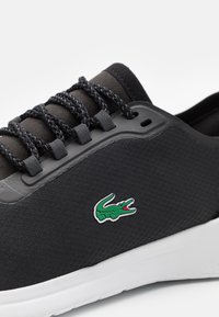 Sneaker noir texturé avec des lacets noirs tressés, semelle blanche et petit logo de crocodile vert sur le côté.