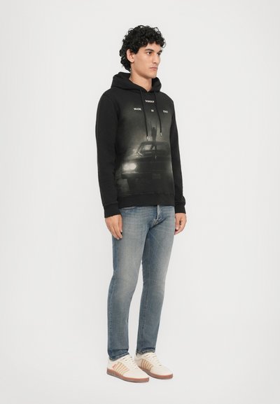 Zwarte hoodie met een grafische print van een auto, verstelbare trekkoorden, gecombineerd met lichtblauwe spijkerbroek en beige sneakers met bruine accenten.