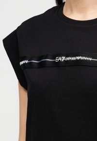 Μαύρο βαμβακερό T-shirt με κοντά μανίκια, διακοσμημένο με οριζόντια μαύρη κορδέλα στο στήθος με λευκό κείμενο της μάρκας "EA7 EMPORIO ARMANI."