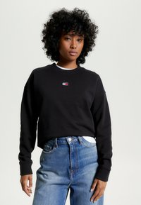 Felpa nera in cotone con colletto rotondo e polsini a coste, caratterizzata da un piccolo logo multicolore sul petto. Abbinata a jeans in denim azzurro chiaro.