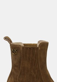 Stivale alla caviglia in suede marrone con cuciture verticali, un emblema decorativo dorato e un'apposita linguetta laterale per una facile calzata.