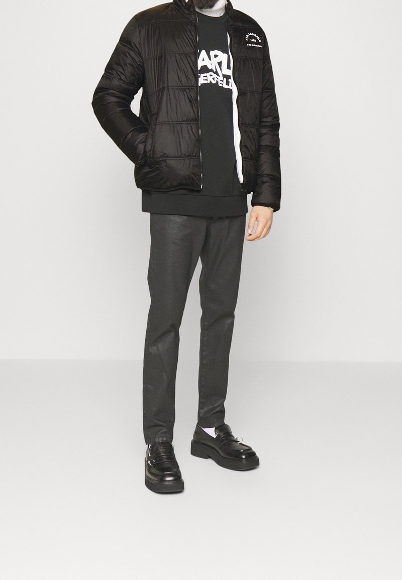 Svart pufferjacka med quiltad design, bärs över en svart sweatshirt med vit text, i kombination med mörka byxor och svarta plattformsskor.