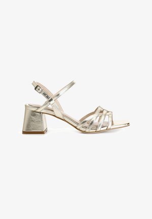 Metallic gold heeled sandals featuring crossed straps, ankle buckle, and a block heel. Smooth texture with a shiny finish.