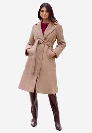 Trench coat beige in misto lana con vita con cintura, larghe rever, tasche laterali e orlo fino al ginocchio. Indossato su un maglione bordeaux e stivali alti marroni.