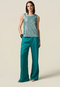 Paljetterlös topp i ljus teal, matchad med vida teal-byxor. Notabla detaljer inkluderar ett svart geometriskt armband och långa örhängen.