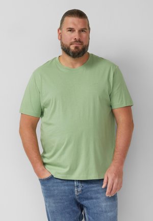 Mann mit Bart, der ein hellgrünes kurzärmeliges T-Shirt und blaue Jeans trägt, vor einem einfarbigen hellgrauen Hintergrund stehend.